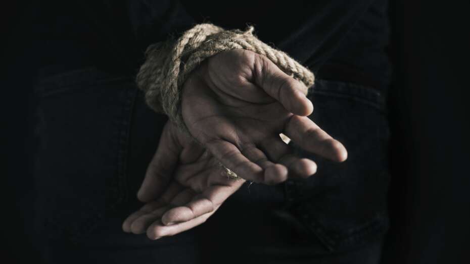 closeup rear view of a man with his hands tied behind his back with rope, against a black background